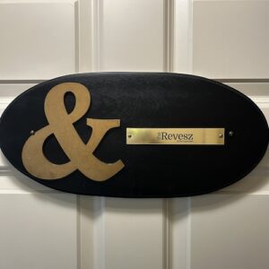 An oval, black room plaque. On the left, there is a gold ampersand. On the right, a gold metal plaque with black writing that reads: "The Revesz at the Ampersand."