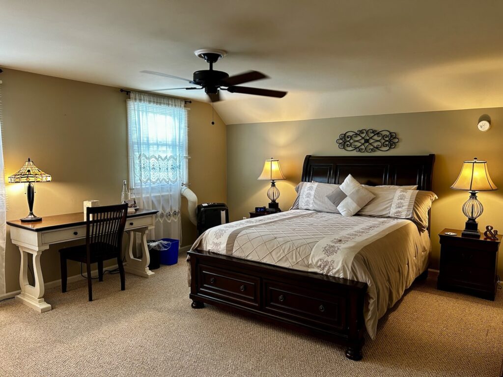 The Costello Room. There is a Queen Bed in the center with two, dark-wood nightstands with lamps on either side. To the left of the bed is a desk and bright windows.