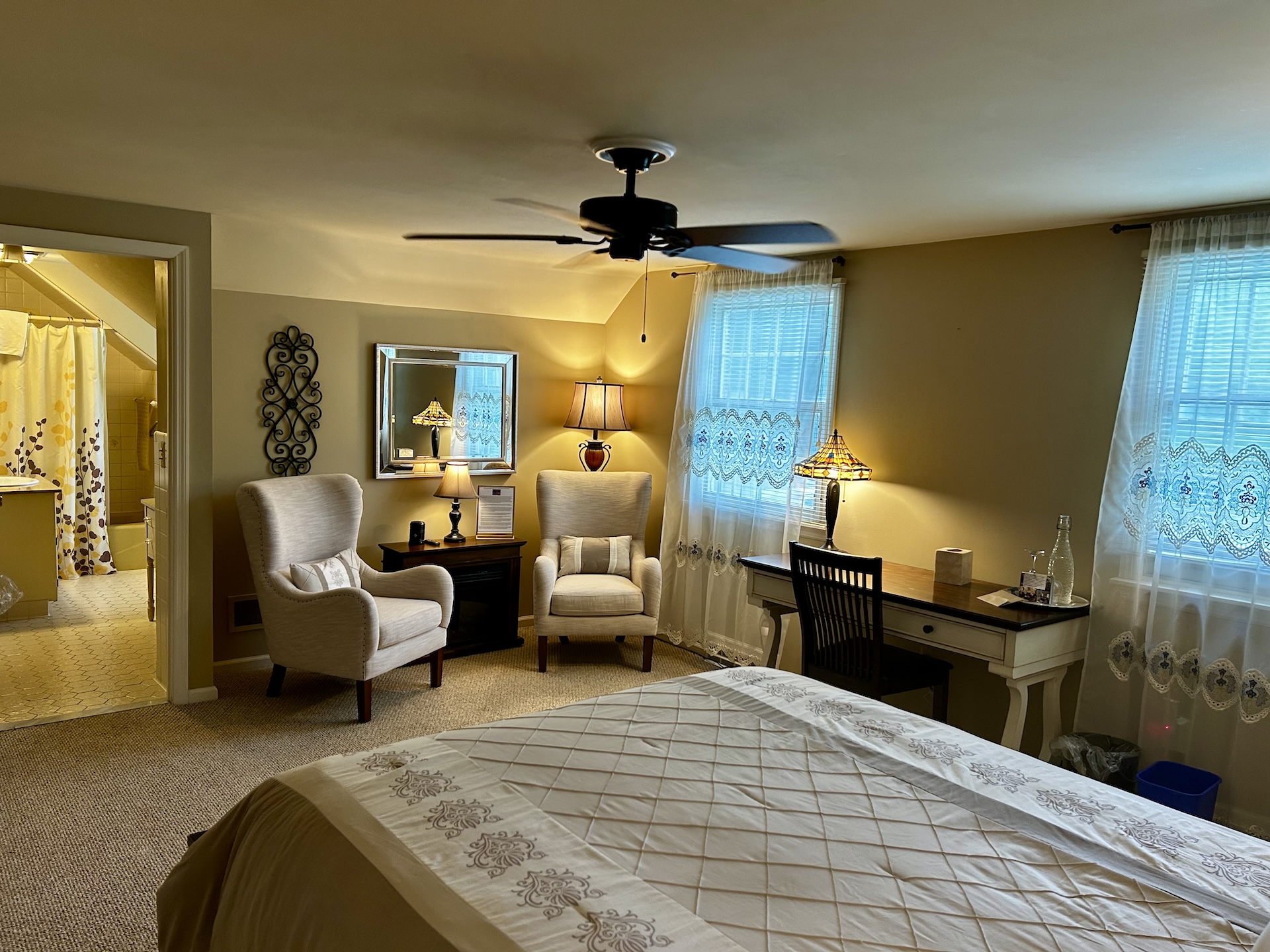 The Costello Room from the view of the queen bed. There is a dark wood desk to the right. On the wall across from the bed is two light-colored, comfy chairs with an electric fireplace between them. To the left is the door to the en-suite bathroom.