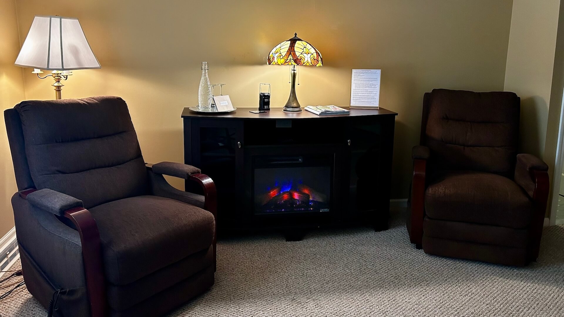 The Coulther Room Seating Area. Two big brown chairs with a table and electric fireplace in between them.
