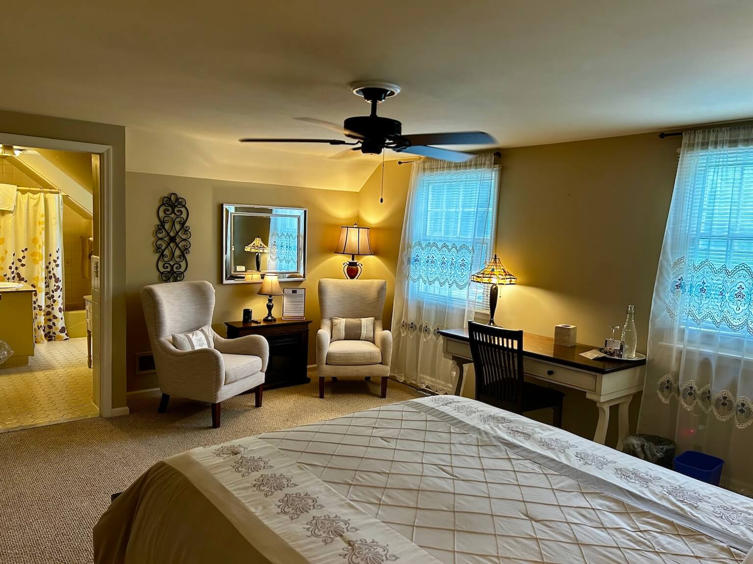 The Costello Room from the view of the Queen bed. There is a dark wood desk to the right. On the wall across from the bed is two light-colored, comfy chairs with an electric fireplace between them. To the left is the door to the en-suite bathroom.